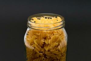 Uncooked Fusilli Pasta in Glass Jar on Black Background. Raw and Dry Macaroni. Unhealthy and Fat Food. Italian Culture photo