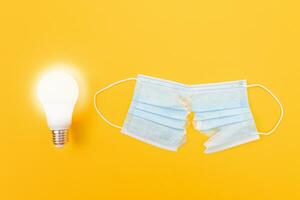Glowing Power Saving Lamp with Torn Medical Face Mask Lying on Yellow Background Anti Mask Concept photo
