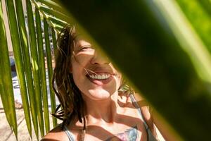 sonriente mujer tomando selfie debajo palma hoja en tropical playa. veraniego vacaciones momento. foto