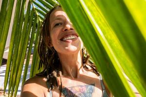 sonriente mujer tomando selfie debajo palma hoja en tropical playa. veraniego vacaciones momento. foto