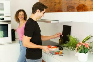 Smiling young man with woman opening wine bottle and holding salad in kitchen at home photo