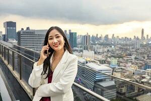 Asian business woman in formal suit is phone calling customer while standing outside the skyscraper building for marketing, connection, corporate work, real estate, housing and urban development photo