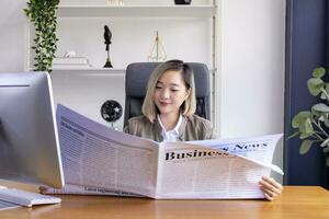 Asian businesswoman working in office using computer while reading newspaper for updating social trend with casual formal wear for business employee concept photo