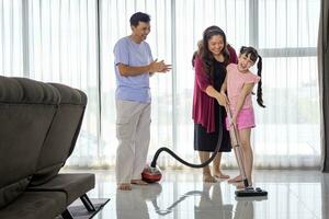 asiático familia con padre, madre y hija ayuda cada otro a limpieza casa utilizando vacío máquina para diario rutina quehaceres y limpieza interna concepto foto