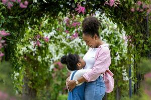 African mother and her daughter hugging each other while happily walking under the blossom trellis arch in the public park during summer for family love and care in mother's day celebration photo