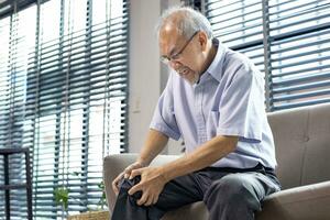 Senior asian man suffering from knee osteoarthritis symptom whiling sitting on the couch at home with copy space for medical surgery treatment and physical therapy photo