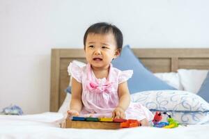 adorable pequeño asiático bebé niñito es sentado en el cama llorando después jugando con su de madera melodía juguete para preescolar aprendizaje y crecimiento desarrollo concepto foto