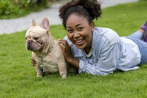 African American woman is playing with her french bulldog puppy while lying down in the grass lawn after having morning exercise in public park photo
