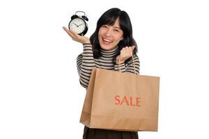 retrato de contento joven asiático mujer con suéter camisa participación alarma reloj y compras papel espalda aislado en blanco antecedentes foto