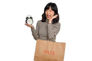 Portrait of excited young Asian woman with sweater shirt holding alarm clock and shopping paper bag isolated on white background. last deals shocked price shopping concept photo