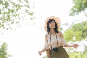 retrato de asiático joven mujer viajero con Costura sombrero y cesta y un cámara en verde público parque naturaleza antecedentes. viaje viaje estilo de vida, mundo viaje explorador o Asia verano turismo concepto. foto