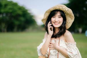 Portrait of asian young woman traveler with weaving hat, basket, mobile phone and camera on green public park background. Journey trip lifestyle, world travel explorer or Asia summer tourism concept. photo