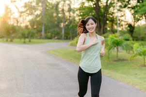 ajuste asiático joven mujer trotar en parque sonriente contento corriendo y disfrutando un sano al aire libre estilo de vida. hembra persona que practica jogging. aptitud corredor niña en público parque. sano estilo de vida y bienestar siendo concepto foto