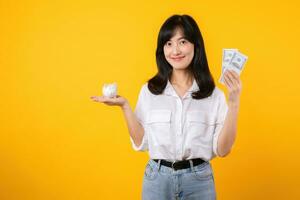 Portrait beautiful young business asian woman wearing white shirt and denim jean with a lot of cash money and piggy bank isolated on yellow background. Wealth money saving, finance investment concept. photo