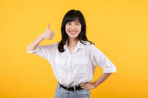 Young asian business woman, smiling while showing thumbs up gesture, recommending product, praise, standing over yellow studio background. Businesswoman confident hand gesture concept. photo