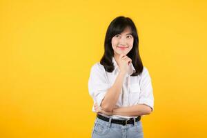 Thoughtful Asian woman keeps hand on chin looks pensively above wearing white shirt and jean plants poses against yellow background blank copy space for your advertising content thinks about future. photo