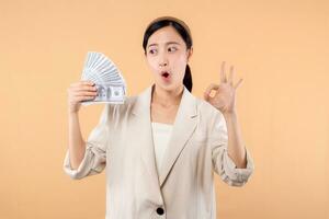 portrait of happy successful confident young asian business woman wearing white jacket holding cash money dollars standing over beige background. millionaire business, shopping concept. photo