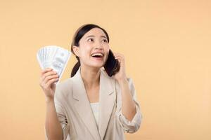 portrait of happy successful confident young asian business woman wearing white jacket holding smartphone and cash money dollar standing over beige background. millionaire business, shopping concept. photo