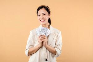 portrait of happy successful confident young asian business woman wearing white jacket holding cash money dollars standing over beige background. millionaire business, shopping concept. photo