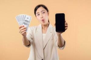 portrait of happy successful confident young asian business woman wearing white jacket holding smartphone and cash money dollar standing over beige background. millionaire business, shopping concept. photo