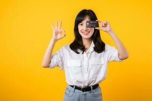 asiático joven mujer participación crédito tarjeta en frente de uno ojo y demostración bueno mano gesto con contento sonrisa aislado en amarillo antecedentes. pago compras en línea concepto. foto