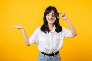 Asian young woman holding credit card in front of one eye and showing pointing hand gesture to free copy space with happy smile isolated on yellow background. Payment shopping online concept. photo