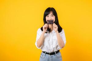 asiático joven mujer participación crédito tarjeta en frente de un boca con contento sonrisa aislado en amarillo antecedentes. pago compras en línea concepto. foto