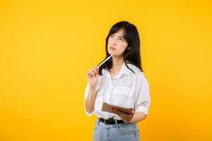 Doubtful Asian young adult business woman working on digital tablet and looking away thinking of solution isolated on yellow background, considering offer, making decision or feeling doubt concept. photo