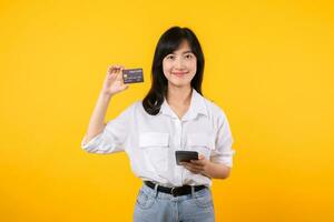 retrato asiático joven mujer vistiendo blanco camisa y mezclilla vaquero con contento sonrisa participación crédito tarjeta a pago compras en línea en teléfono inteligente aislado en amarillo antecedentes. compras en línea concepto. foto