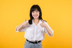 Young asian business woman, smiling while showing thumbs up gesture, recommending product, praise, standing over yellow studio background. Businesswoman confident hand gesture concept. photo