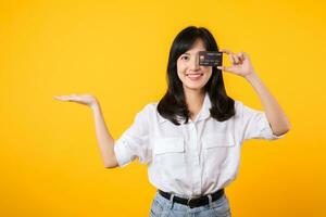 Asian young woman holding credit card in front of one eye and showing pointing hand gesture to free copy space with happy smile isolated on yellow background. Payment shopping online concept. photo