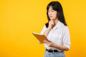 Doubtful Asian young adult business woman working on digital tablet and looking away thinking of solution isolated on yellow background, considering offer, making decision or feeling doubt concept. photo