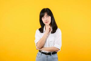 Thoughtful Asian woman keeps hand on chin looks pensively above wearing white shirt and jean plants poses against yellow background blank copy space for your advertising content thinks about future. photo