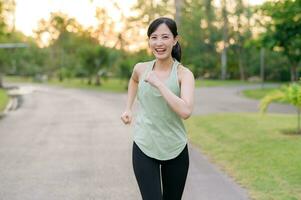 ajuste asiático joven mujer trotar en parque sonriente contento corriendo y disfrutando un sano al aire libre estilo de vida. hembra persona que practica jogging. aptitud corredor niña en público parque. sano estilo de vida y bienestar siendo concepto foto