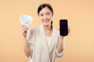 portrait of happy successful confident young asian business woman wearing white jacket holding smartphone and cash money dollar standing over beige background. millionaire business, shopping concept. photo