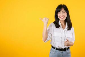 Happy young asian woman wearing white shirt and denim jean plants pointing thumb finger away on copy space isolated on yellow background. helps pick best choice, recommends product concept. photo