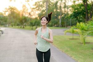 ajuste asiático joven mujer trotar en parque sonriente contento corriendo y disfrutando un sano al aire libre estilo de vida. hembra persona que practica jogging. aptitud corredor niña en público parque. sano estilo de vida y bienestar siendo concepto foto