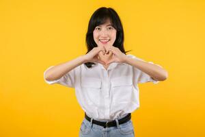 Young female healthcare. Asian woman wearing white shirt feels happy and romantic shapes heart gesture expresses tender feeling poses isolated on yellow background. People affection and care concept. photo