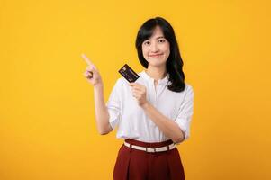 Portrait beautiful young asian woman enterpriser happy smile wearing white shirt and red plants pointing finger gesture to free copy space and holding credit card isolation on yellow background. photo