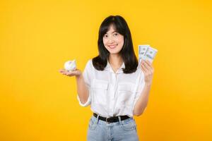 retrato hermosa joven negocio asiático mujer vistiendo blanco camisa y mezclilla vaquero con un lote de efectivo dinero y cerdito banco aislado en amarillo antecedentes. riqueza dinero ahorro, Finanzas inversión concepto. foto