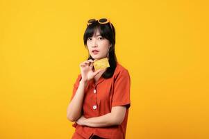 Portrait young beautiful asian woman happy smile dressed in orange clothes showing credit card isolated on yellow background. Pay and purchase shopping payment concept. photo