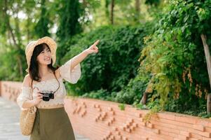 retrato de asiático joven mujer viajero con Costura sombrero y cesta y un cámara en verde público parque naturaleza antecedentes. viaje viaje estilo de vida, mundo viaje explorador o Asia verano turismo concepto. foto