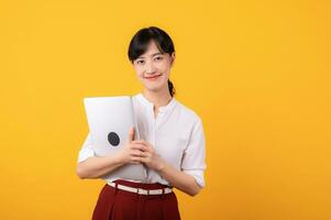 Portrait beautiful young asian woman enterpriser happy smile wearing white shirt and red plants showing confident body language gesture and holding laptop isolation on yellow background. photo