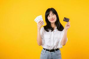 happy successful confident young asian woman happy smile wearing white shirt and denim jean holding cash money and credit card standing over yellow background. millionaire business, shopping concept. photo