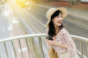 young asian woman traveler with weaving basket using mobile phone and standing on overpass with railway background. Journey trip lifestyle, world travel explorer or Asia summer tourism concept. photo