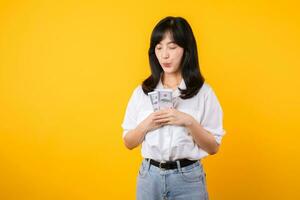 contento joven asiático mujer de negocios vistiendo blanco camisa y mezclilla vaquero participación dinero en efectivo, abrazando dolares dinero y sonriente, en pie terminado amarillo antecedentes. amor a ser Rico concepto. foto