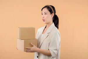 portrait of happy successful confident young asian business woman wearing white jacket holding parcel box isolated on beige studio background, Delivery courier and shipping service concept. photo
