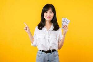 Young happy woman of Asian ethnicity wear white shirt and denim jean holding cash money in dollar and pointing finger to free copy space against yellow background. Investment and financial concept. photo