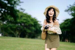 Portrait of asian young woman traveler with weaving hat and basket and a camera on green public park nature background. Journey trip lifestyle, world travel explorer or Asia summer tourism concept. photo