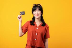 Portrait young beautiful asian woman happy smile dressed in orange clothes showing credit card isolated on yellow background. Pay and purchase shopping payment concept. photo